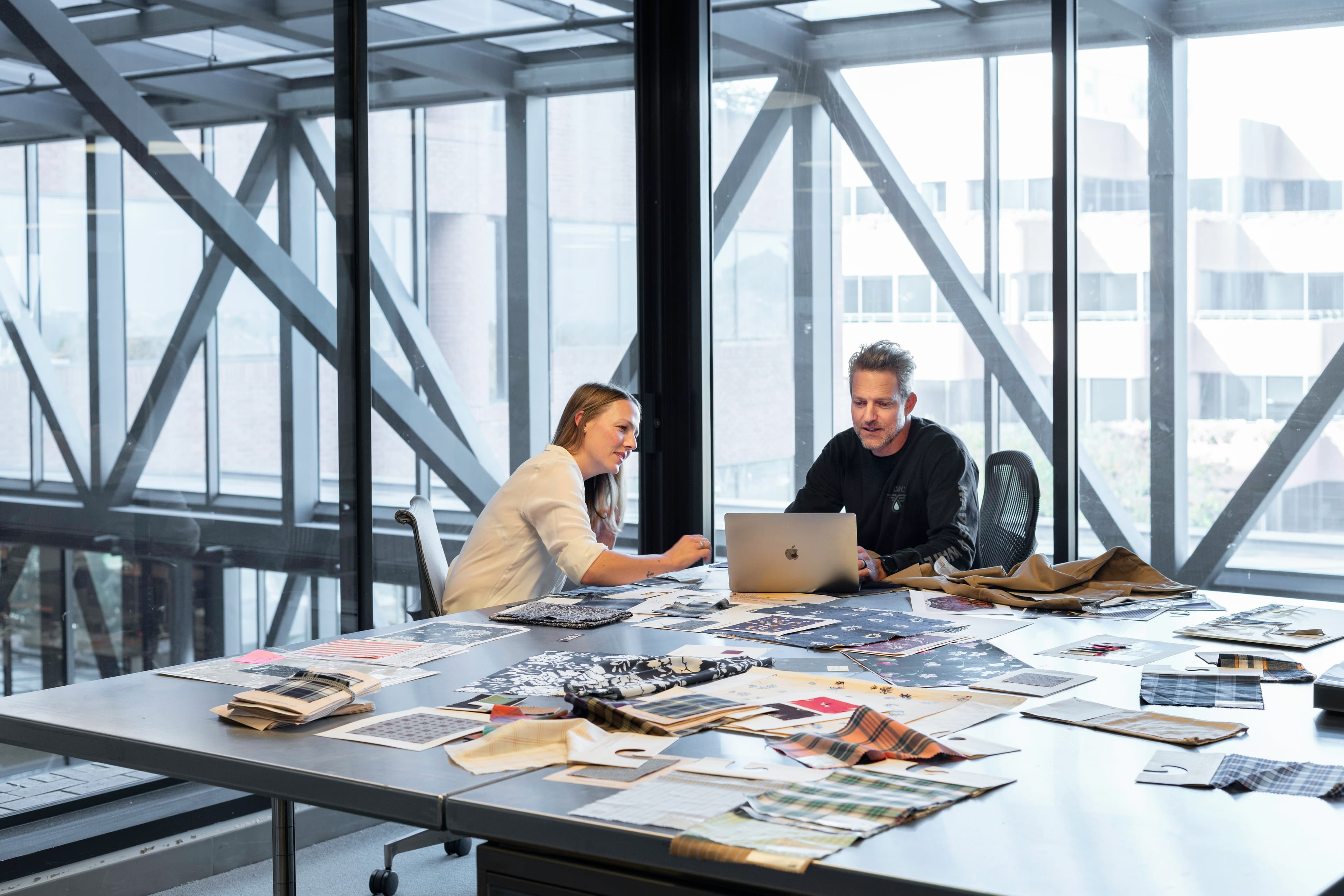 Two people reviewing work together over a laptop at a long table by a window